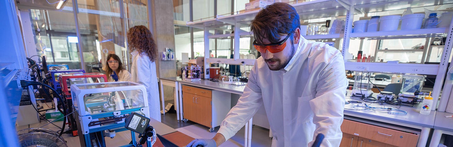 Man in lab coat and safety glasses with 3d printers and two women to the left