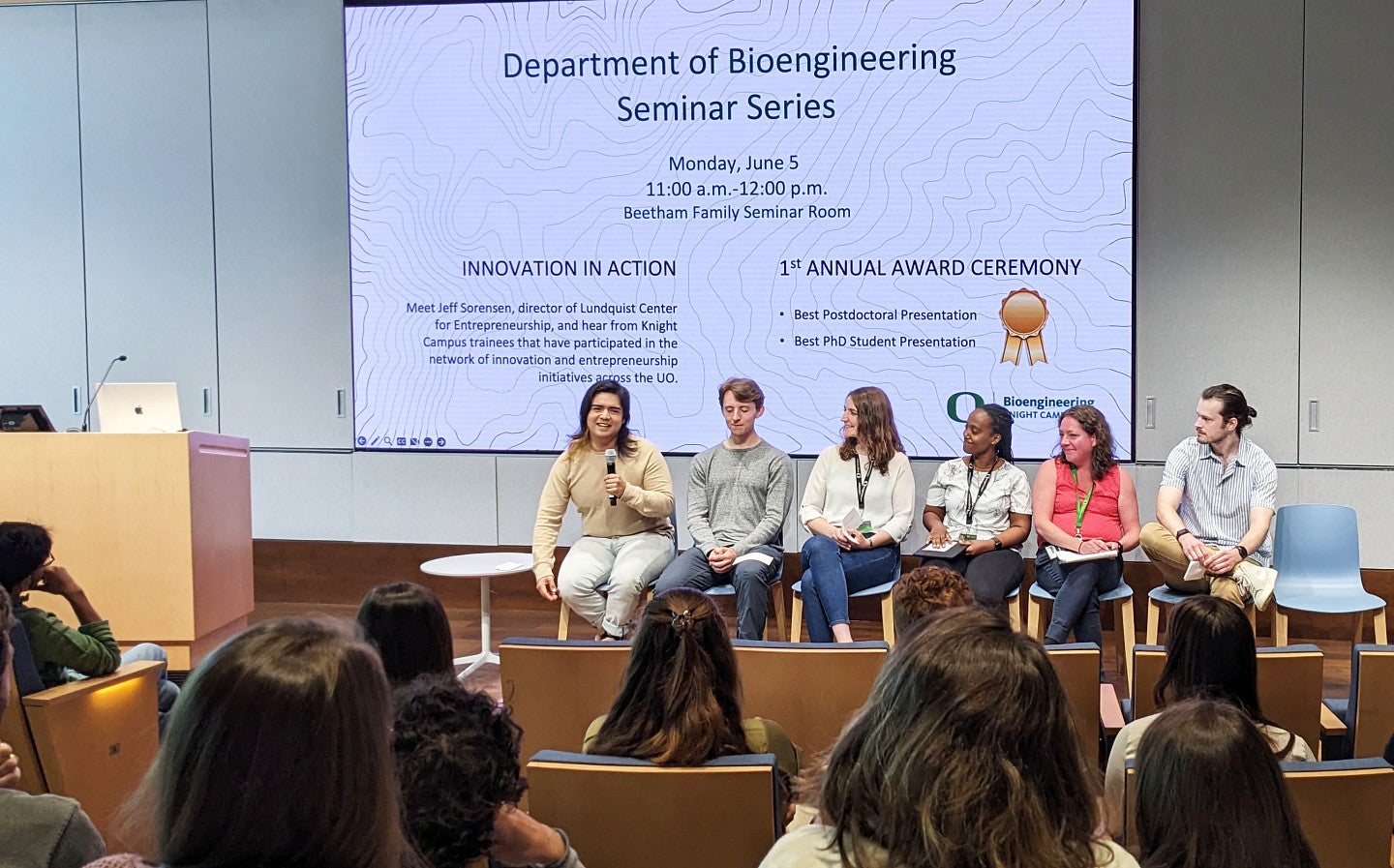 Six adults sit on chairs and speak to an audience