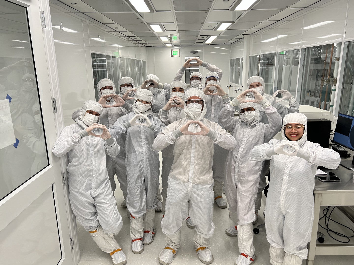 A large group of people in PPE gear pose for a photo with their hands in the shape of an "O" outside a clean room