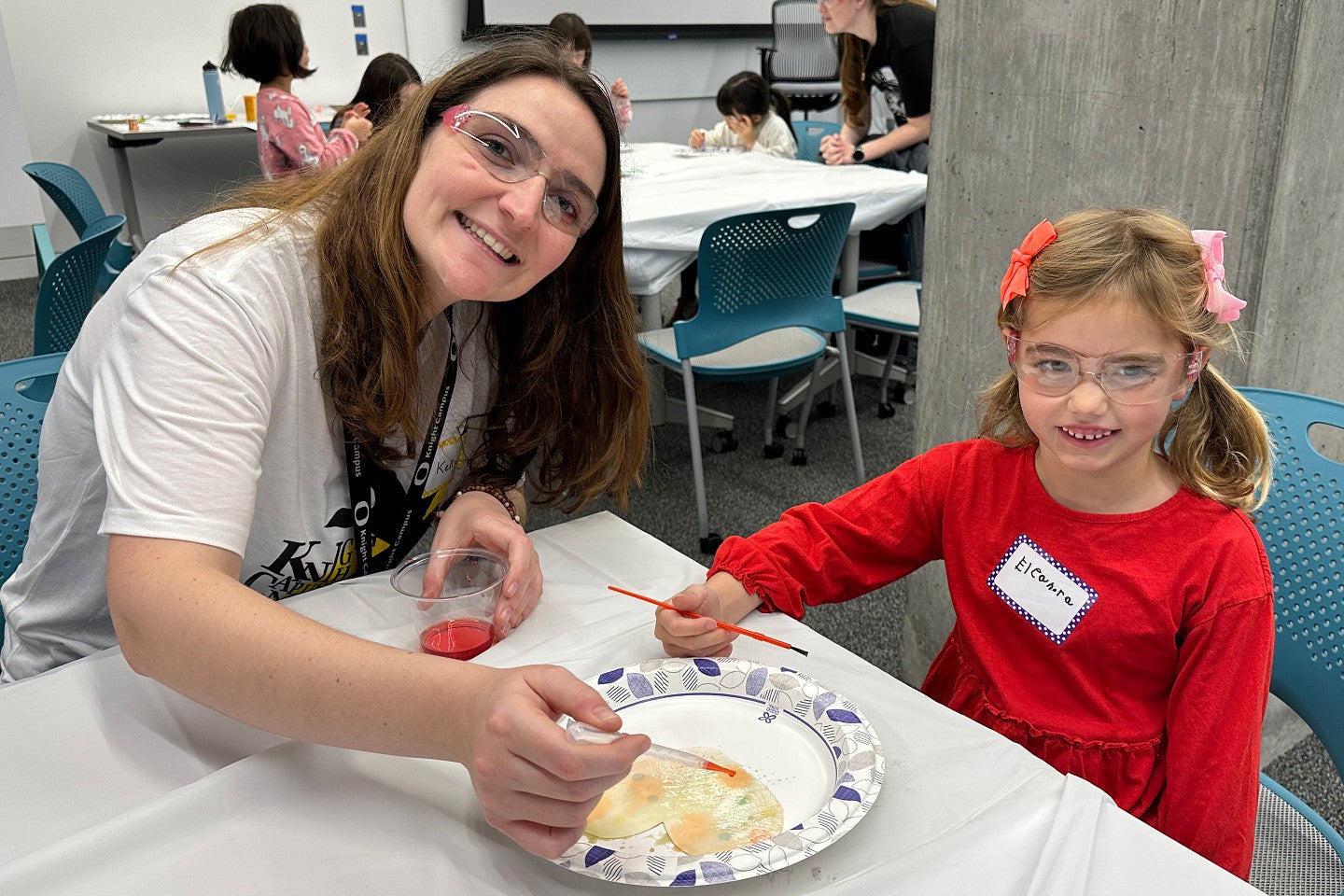 Adult and child painting together at a table during a workshop, with other participants in the background.
