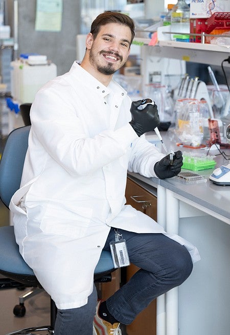 Yan Carlos Pacheco sitting on a stool with a pippette in hand smiling in the lab