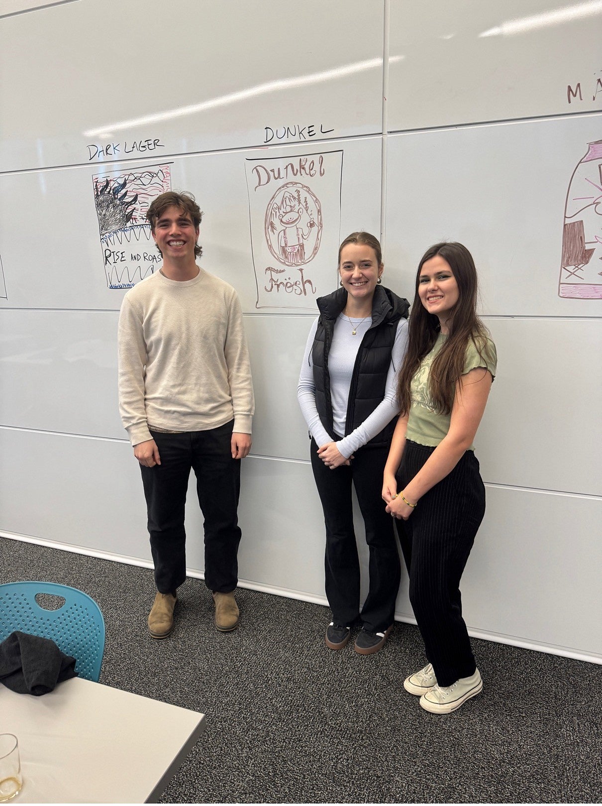 Three people standing in front of white board with drawing of beer can labeled Dunkel Fresh