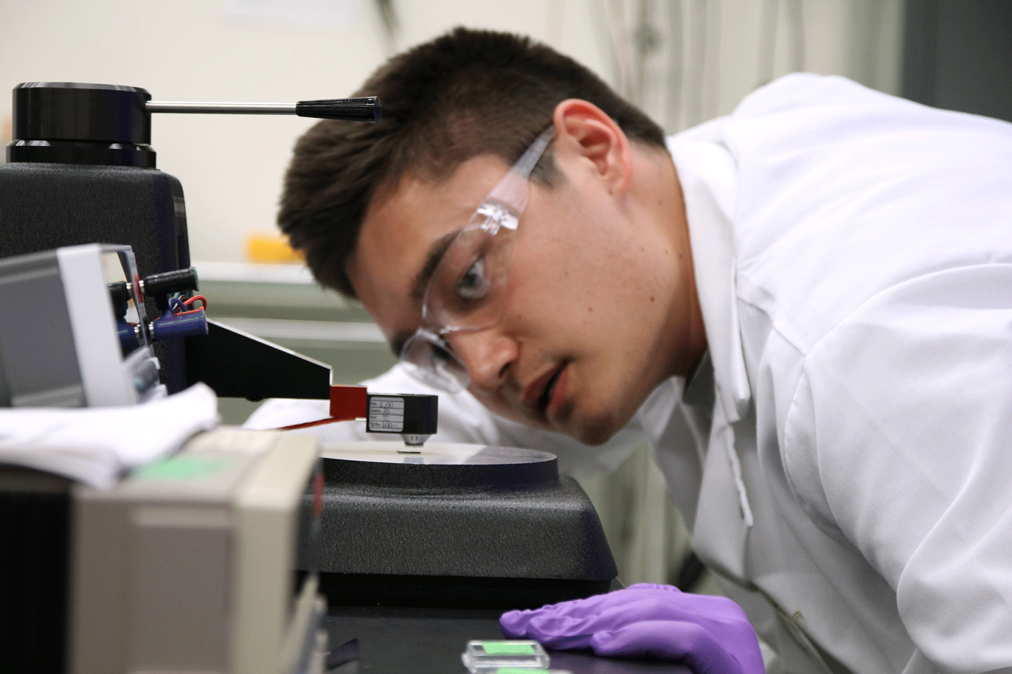Person in white lab coat and safety glasses inspecting semiconductor wafer