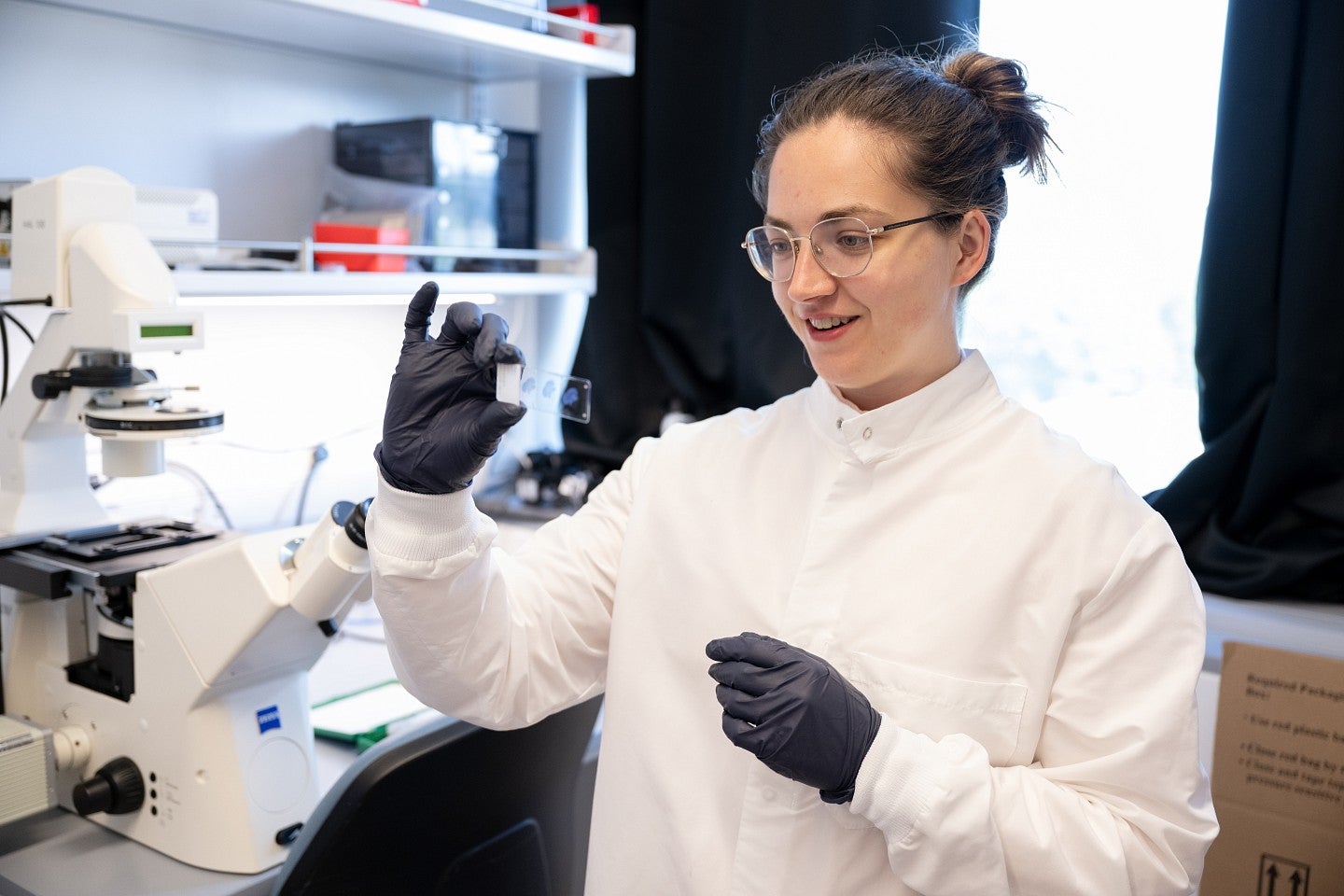 Graduate student, Rose Hulsey-Vincent, at the microscope looking at a slide, in a white lab coat and gloves