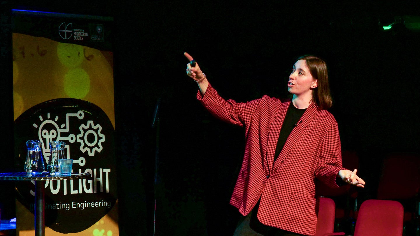 Sara Keller, wearing a large blazer, pointing at a screen, giving an public talk about her research