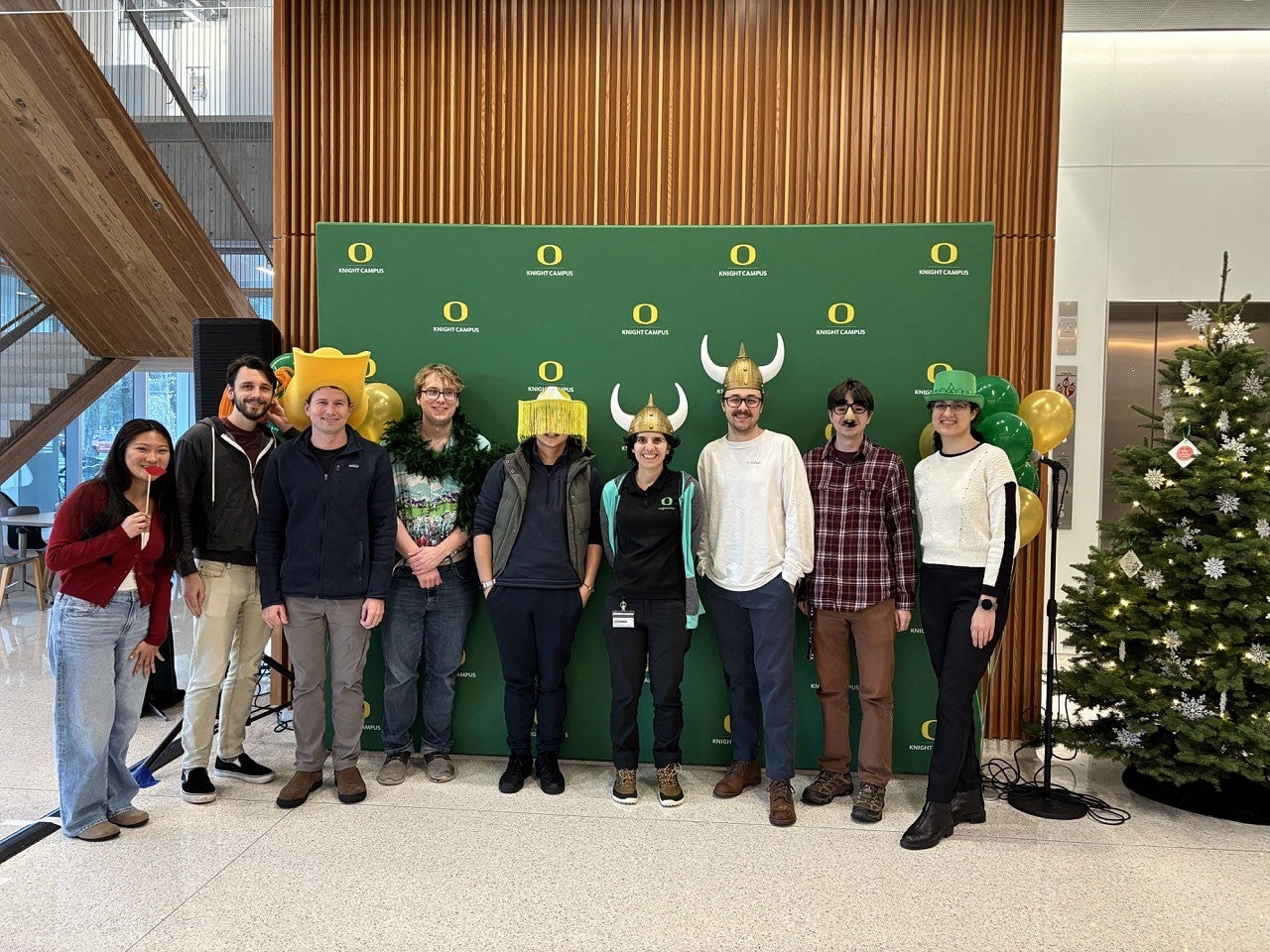 A group of people wearing fun hats and costumes in front of a green pull up banner at the annual holiday party