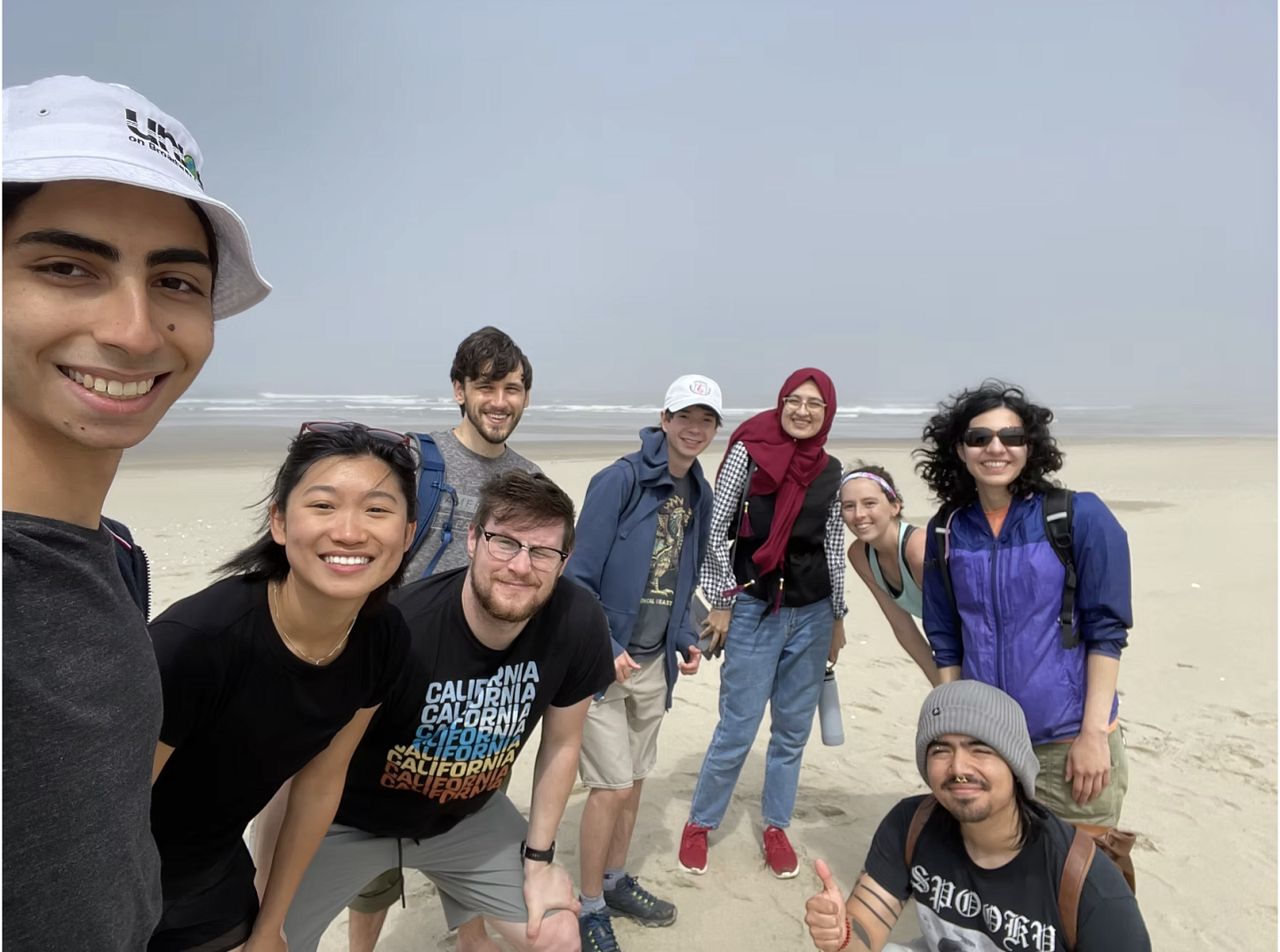Group of people looking at the camera, and smiling. they are on a sandy beach, with the ocean in the background - it is windy and overcast