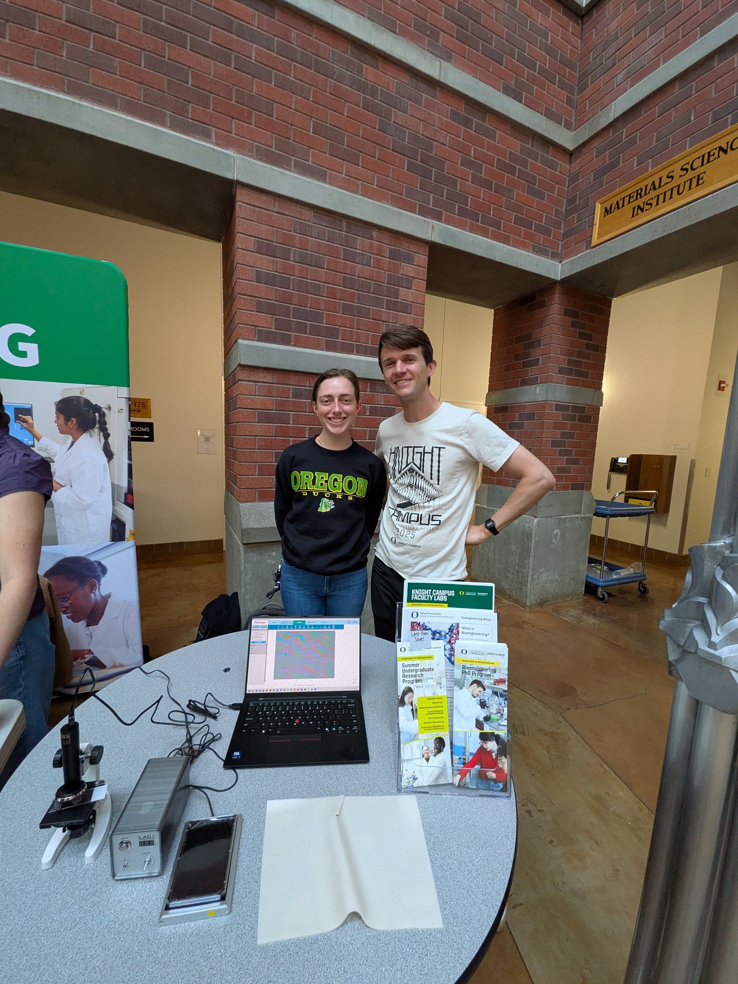 2 people standing in front of a brick wall, standing in front of a table with some flyers and handouts