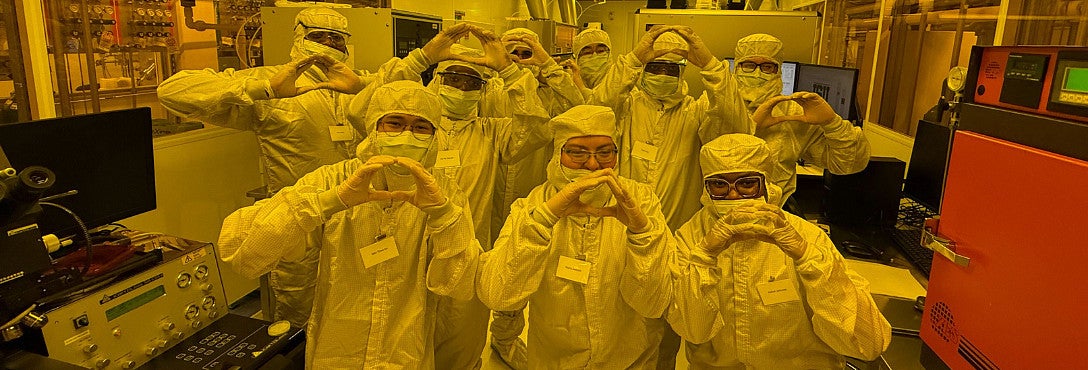 Group of people in anti-contamination suits— bunny suits, hoods, masks, and safety glasses — pose together in a semiconductor cleanroom bathed in yellow lithography-safe lighting, each forming the University of Oregon's "O" hand symbol with their fingers in front of their chest