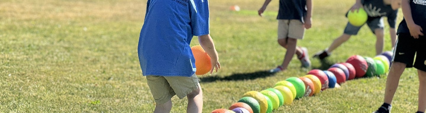 A child in a blue shirt playing a ball game, carefully stepping on colorful balls aligned on a field, with other children in the background.