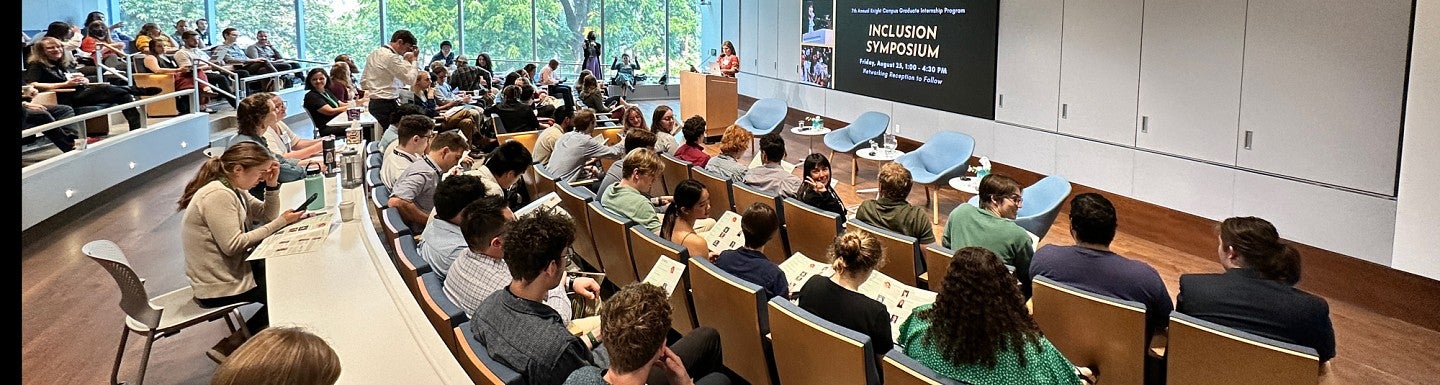 Seminar room with audience in the foreground, speaker at podium in background and slide on screen reading "Inclusion Symposium"