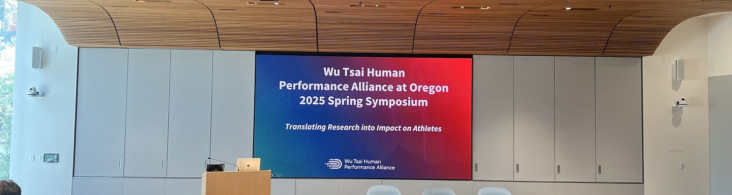 A mostly empty seminar room, with wood ceilings, and a screen displaying 'Wu Tsai Human Performance Alliance at Oregon 2025 Spring Symposium' with a a blue to red gradient
