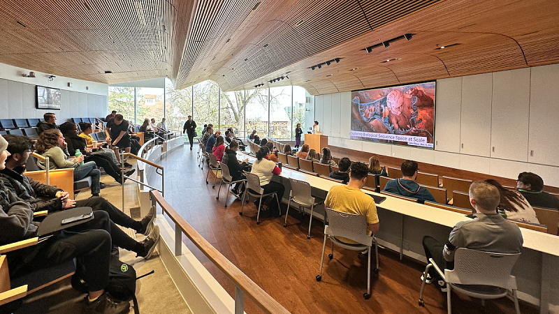 Students and faculty in campus lecture room with speaker on stage and slide on projection screen