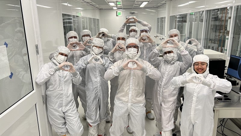 A large group of people in PPE gear pose for a photo with their hands in the shape of an "O" outside a clean room