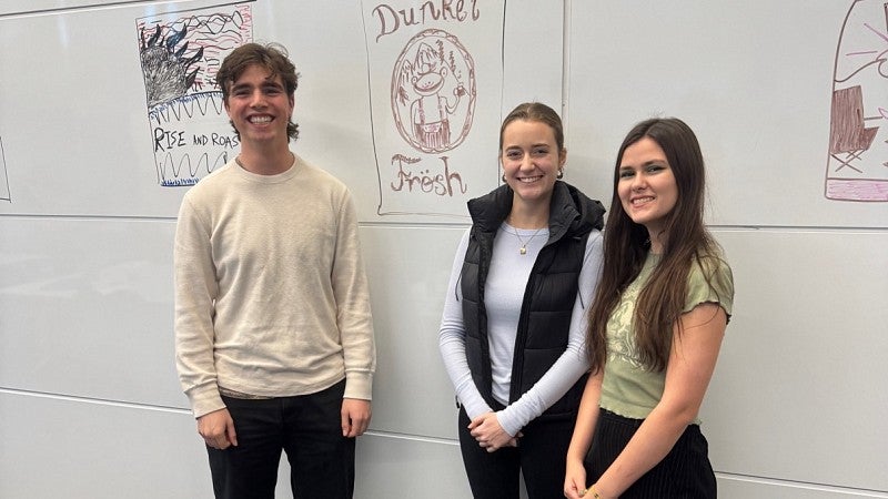 Three people standing in front of white board with drawing of beer can labeled Dunkel Fresh