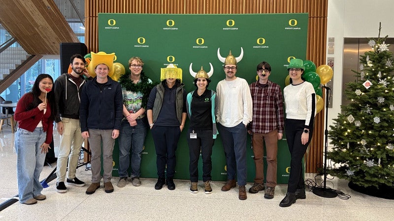 A group of people wearing fun hats and costumes in front of a green pull up banner at the annual holiday party