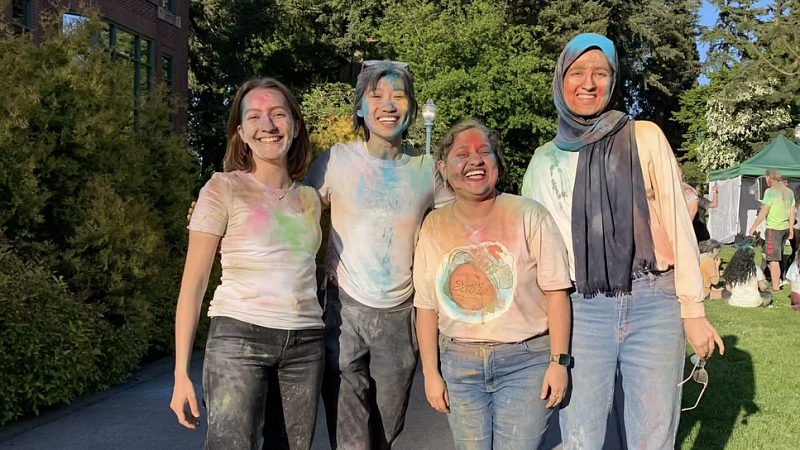 group of students smiling, looking at the camera, wearing white shirts with colored powder over them for a Holi celebration