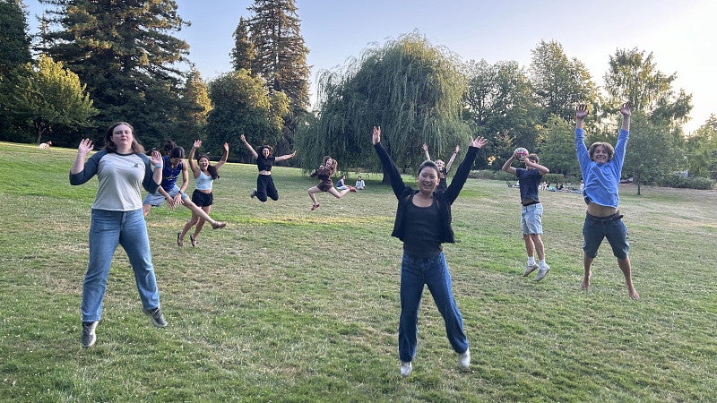 a group of people jumping up, in a scenic park with a blue sky and trees