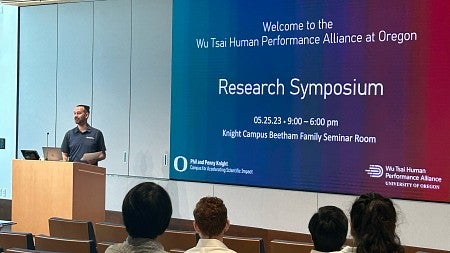 Person at lectern with audience in foreground and slide on screen welcoming people to the Wu Tsai research symposium