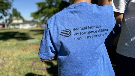 Child in a blue t-shirt with "Wu Tsai Human Performance Alliance UNIVERSITY OF OREGON" text on the back, standing in a sunny park with other partially visible children in the foreground.