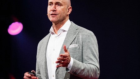 Person on stage with white shirt, gray sportjacked and tan pants with black backdrop and pink lights