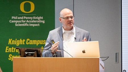Man with glasses, in a white shirt with a grey blazer jacket in front of a green banner, at a podium, actively talking