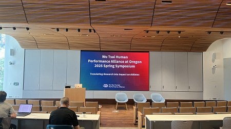 A mostly empty seminar room, with wood ceilings, and a screen displaying 'Wu Tsai Human Performance Alliance at Oregon 2025 Spring Symposium' with a a blue to red gradient