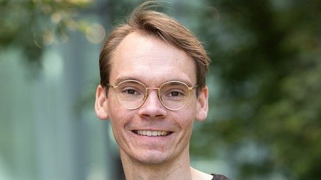Headshot of a man with glasses, in a black shirt, on an outdoor terrace