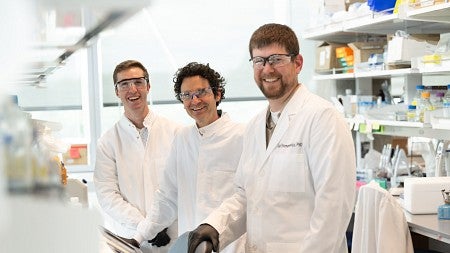 researchers in the Plesa lab, wearing goggles, gloves and labcoats, smiling at the camera. 