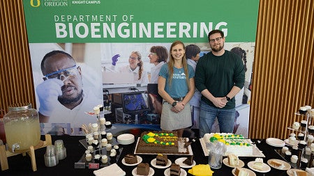 Two people standing in front of a table with sheet cake and deserts, and a bioengineering banner behind them