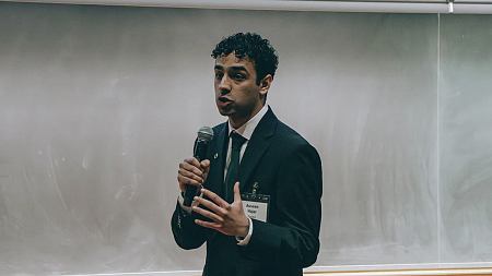 Student in formal attire with microphone in mid speech with chalkboard behind him