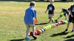 A child in a blue shirt playing a ball game, carefully stepping on colorful balls aligned on a field, with other children in the background.