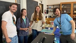 Four individuals in a science laboratory, smiling and standing around a table with experimental equipment. The table holds a structure resembling a bridge made of yellow and orange sticks, and various tools and lab apparatus are visible in the background