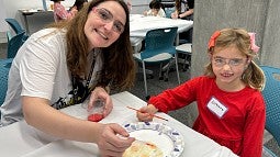 Adult and child painting together at a table during a workshop, with other participants in the background.