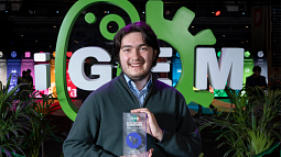 Person standing with trophy in hand before a green and white sculptural backdrop that reads iGEM