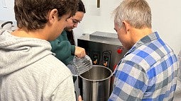 Three people stand around a brewing kettle brewing beer