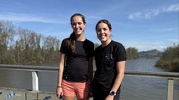 Two women standing on a bridge, with a water and blue skies in the background, wearing running apparel 
