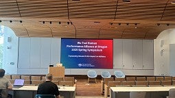 A mostly empty seminar room, with wood ceilings, and a screen displaying 'Wu Tsai Human Performance Alliance at Oregon 2025 Spring Symposium' with a a blue to red gradient