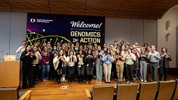 a large group of participants at 2026 genomics in action, in the seminar room, holding the "o"