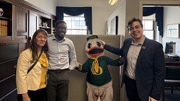 three people standing in front of a desk, at capitol hill, with the oregon duck mascot in the middle