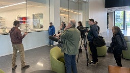 A man in a red hoodie, standing in front of a group of reporters, by a glass wall where you can see into a laboratory space