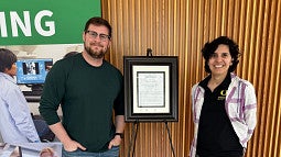 two people standing in front of a framed plaque, with a banner behind them
