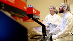 Two people in lab coats and head scarfs in front of fume hoods