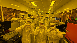 A group of participants wearing full cleanroom gowning — bunny suits, hoods, masks, and safety glasses — pose together in a semiconductor cleanroom bathed in yellow lithography-safe lighting, each forming the University of Oregon's "O" hand symbol with their fingers in front of their chest.