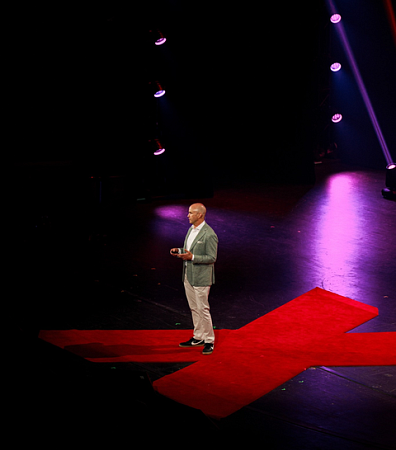 Person in gray sportcoat, white shirt and tan pants on stage at TEDx event standing on red "X" on stage with black backdrop and pink stage lights