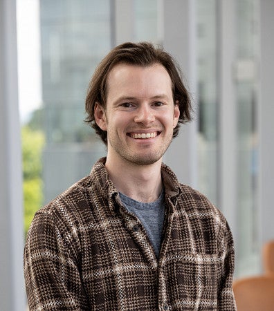 A man with dark, brown hair, smiling for the camera, wearing a brown flannel top, for a headshot