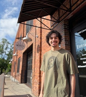 Young man stands in front of brick building with sign that reads "The Wheel"