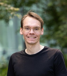 Headshot of a man with glasses, in a black shirt, on an outdoor terrace