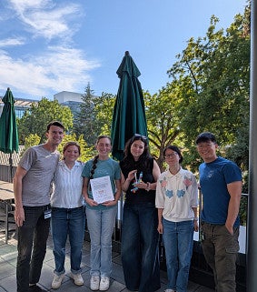 Six people standing on terrace with greenery in backdrop and two members holding certificates